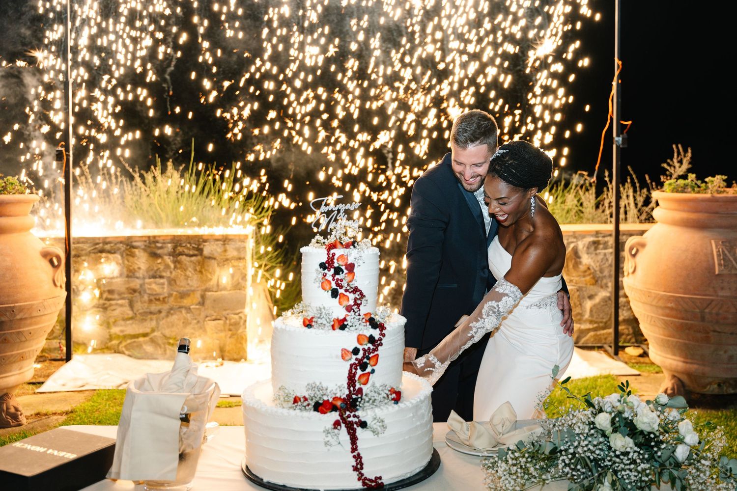 Wedding couple cuts their elegant white tiered cake decorated with red floral accents at an outdoor evening reception.