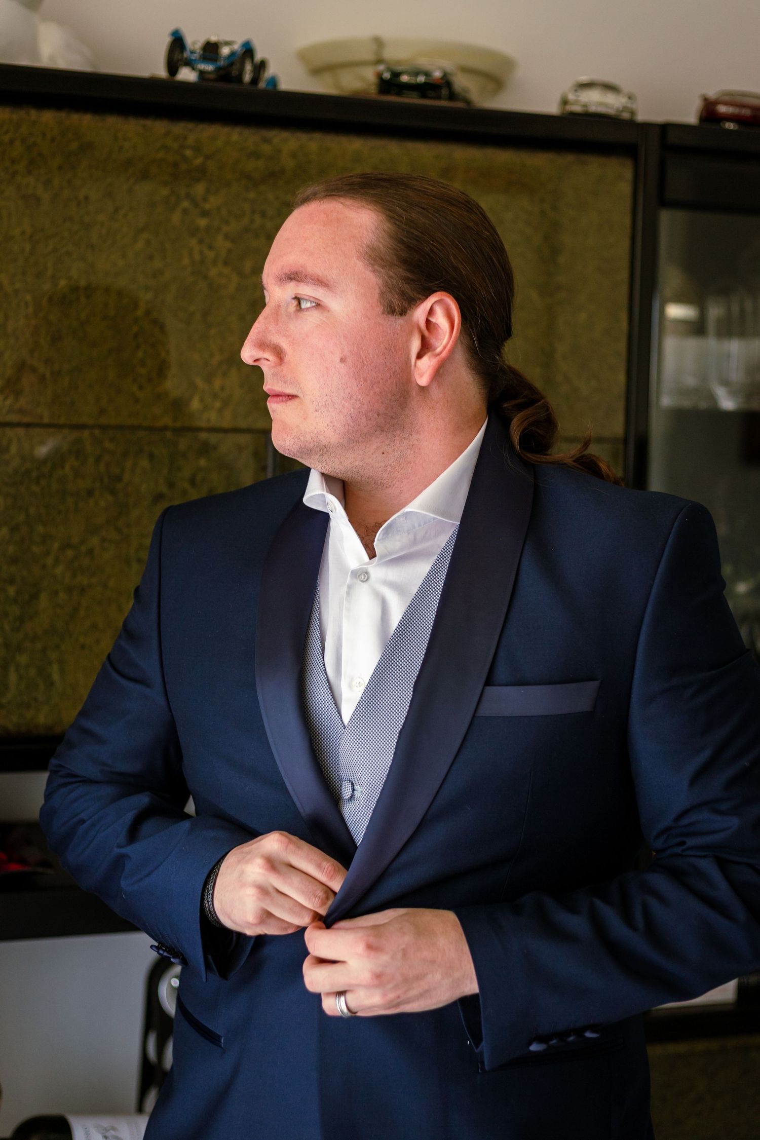 Man in navy blue tuxedo jacket buttoning his suit while looking away, wearing a white shirt and gray vest.