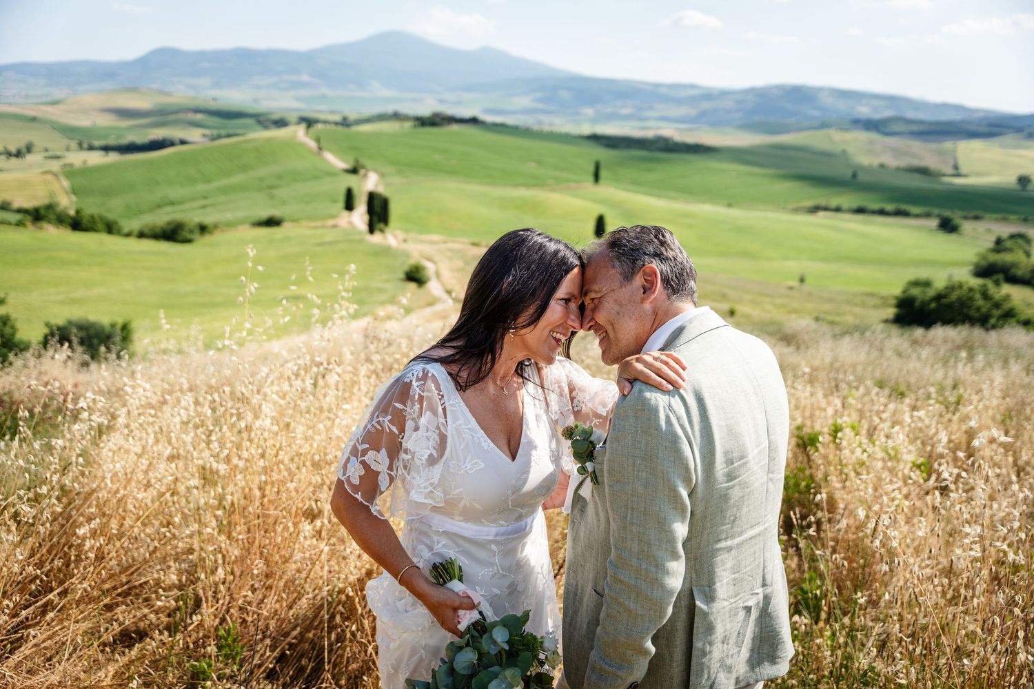 Couple embracing in golden wheat field with rolling green hills and mountains in the background.
