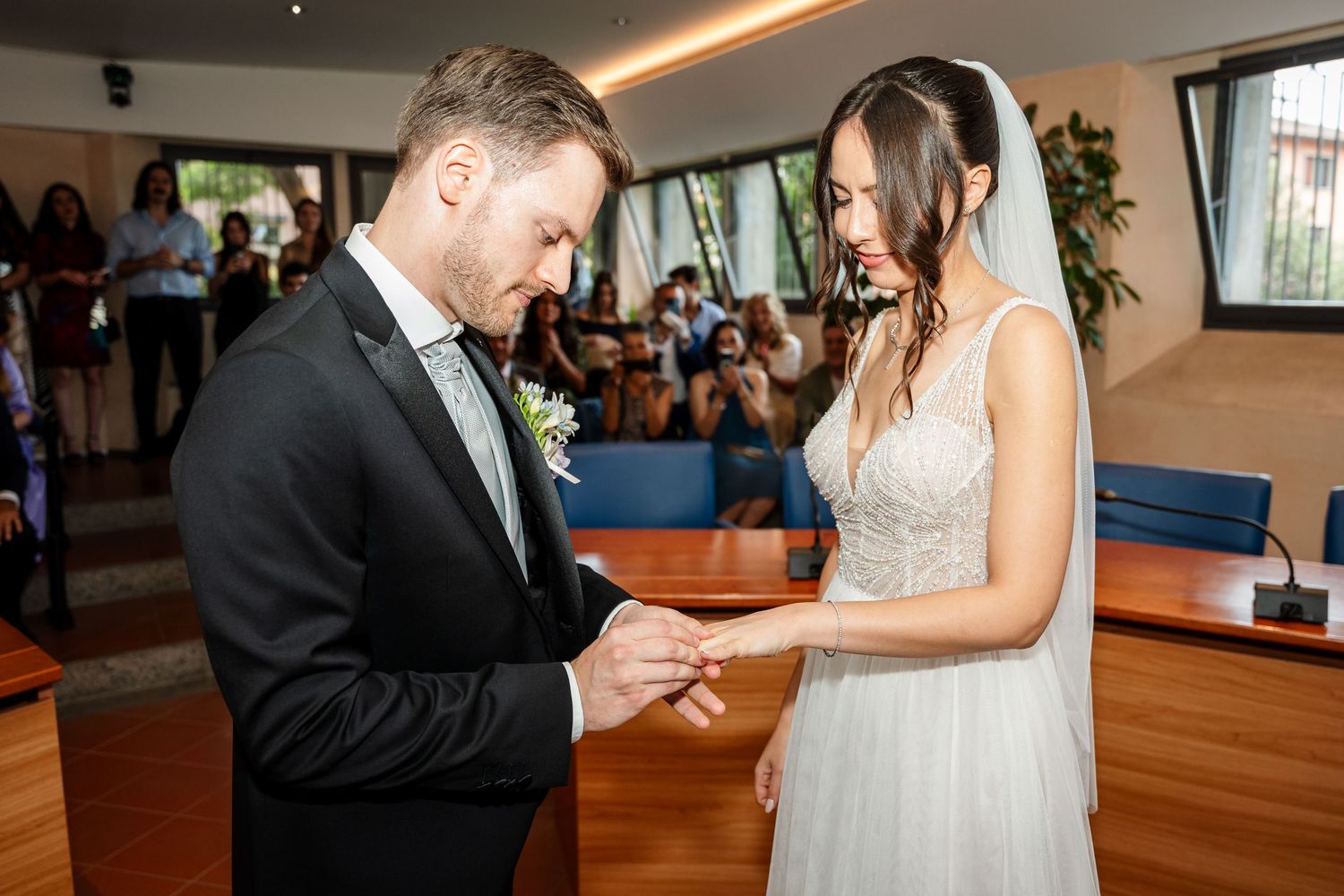 Groom placing wedding ring on brides finger during ceremony with guests.