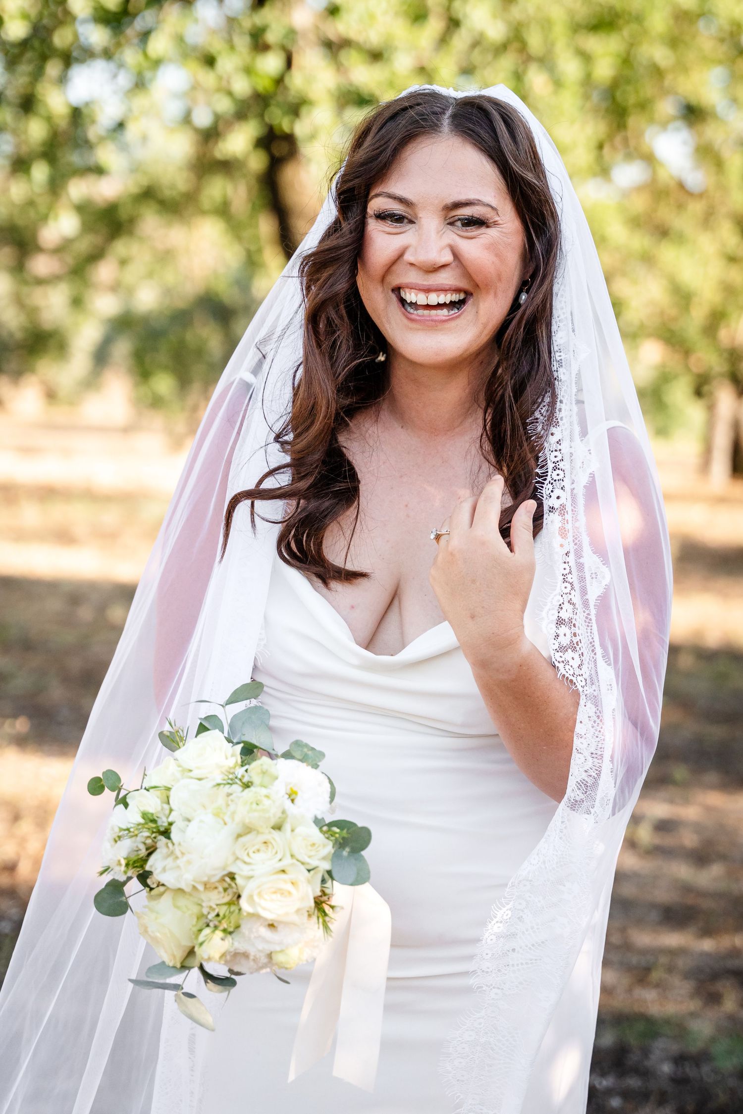 Laughing bride in white satin gown and veil holding white rose bouquet outdoors.