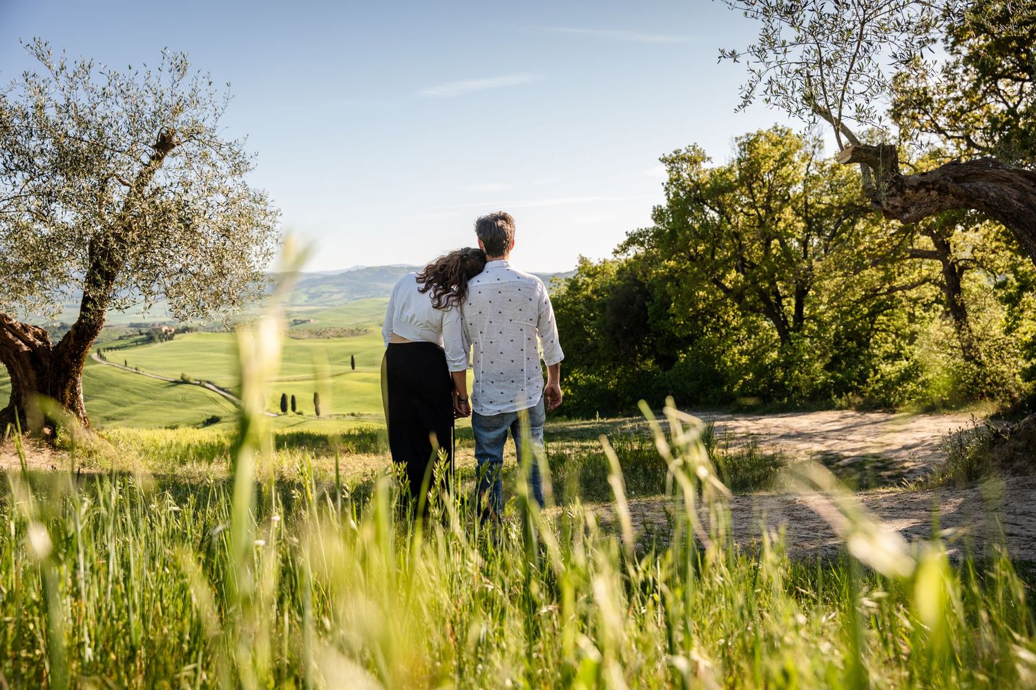 Couple walking hand in hand through lush green Tuscan countryside with rolling hills and olive trees on a sunny day.