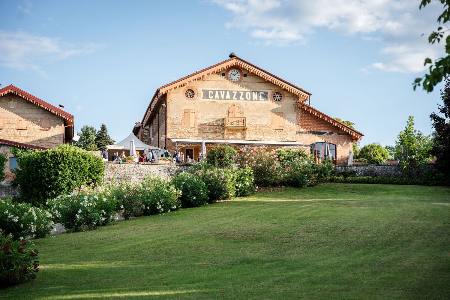 Rustic stone winery building with lush green lawn and colorful gardens under a bright blue sky.
