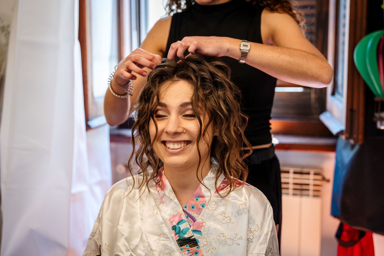 Smiling bride with curly hair getting her hairstyle done by a stylist before her wedding ceremony.