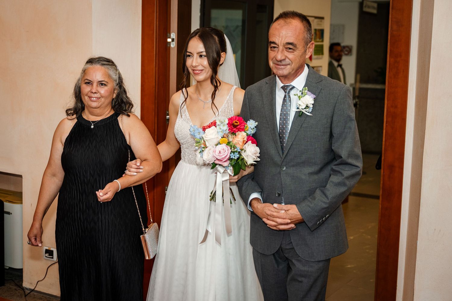 Bride in white gown walks with parents before wedding ceremony.