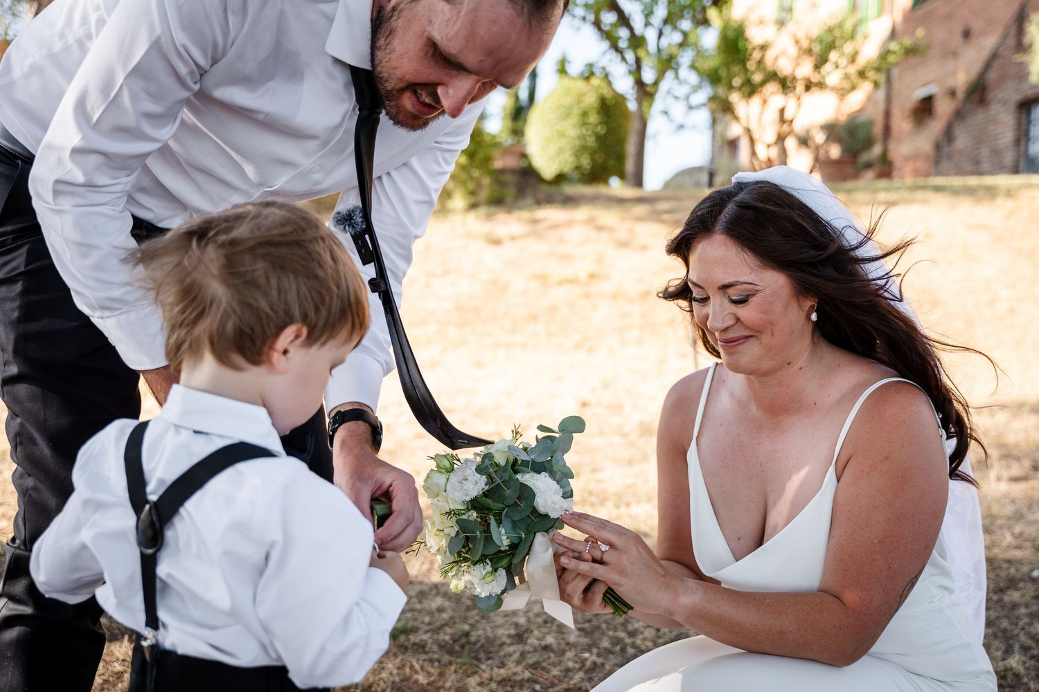 Bride and groom kneel to present bouquet to young ring bearer in suspenders outdoors.
