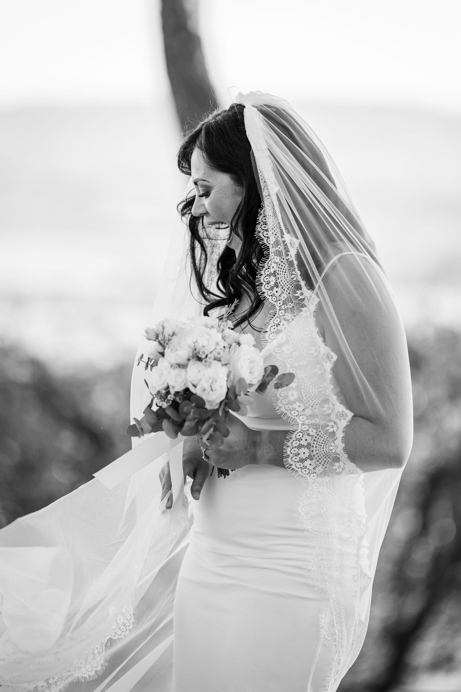 A black and white photo of a bride in a fitted lace gown and veil holding a bouquet outdoors.