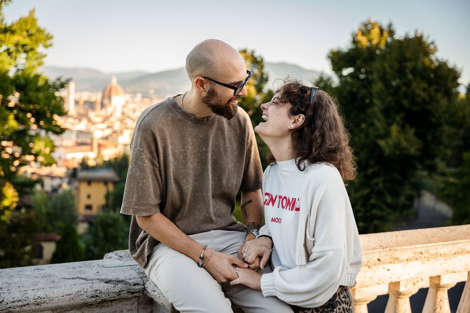 A romantic couple shares tender moments while sitting on a stone wall overlooking a scenic European cityscape at sunset.