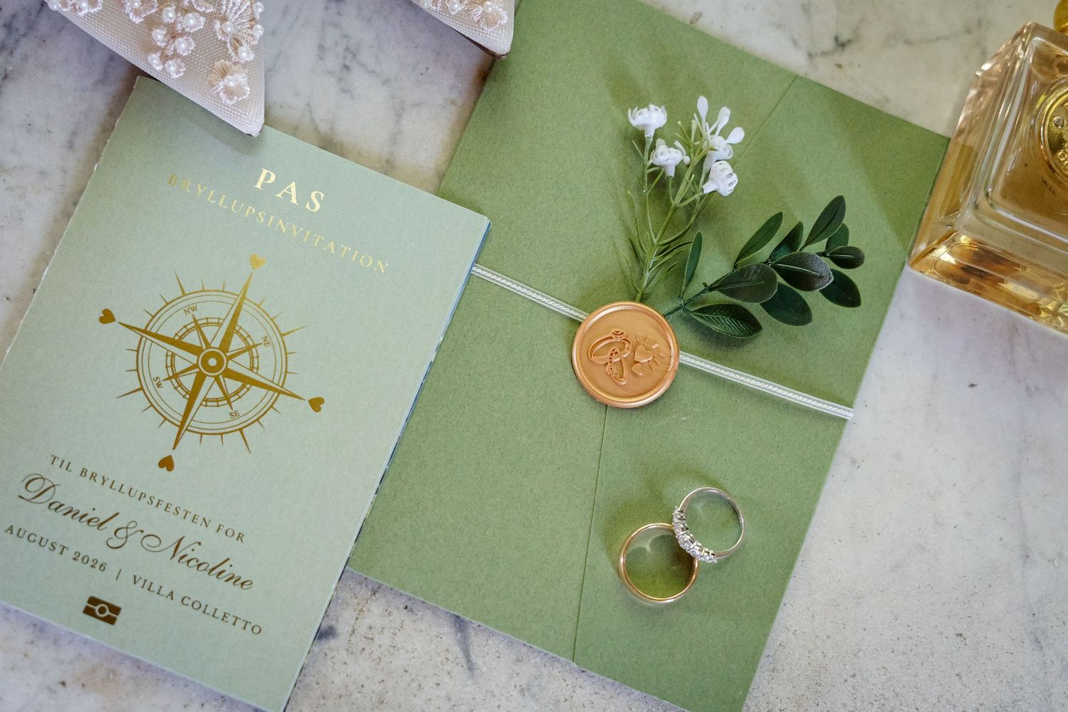 Elegant green wedding invitation with gold wax seal, compass rose design, rings, and white flowers on marble surface.