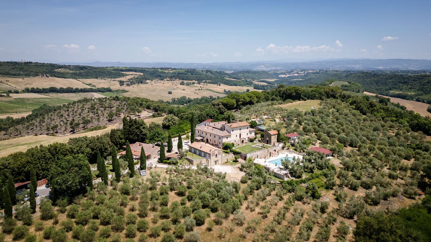 Aerial view of a Tuscan villa surrounded by olive trees and cypress trees with rolling countryside hills in the background.