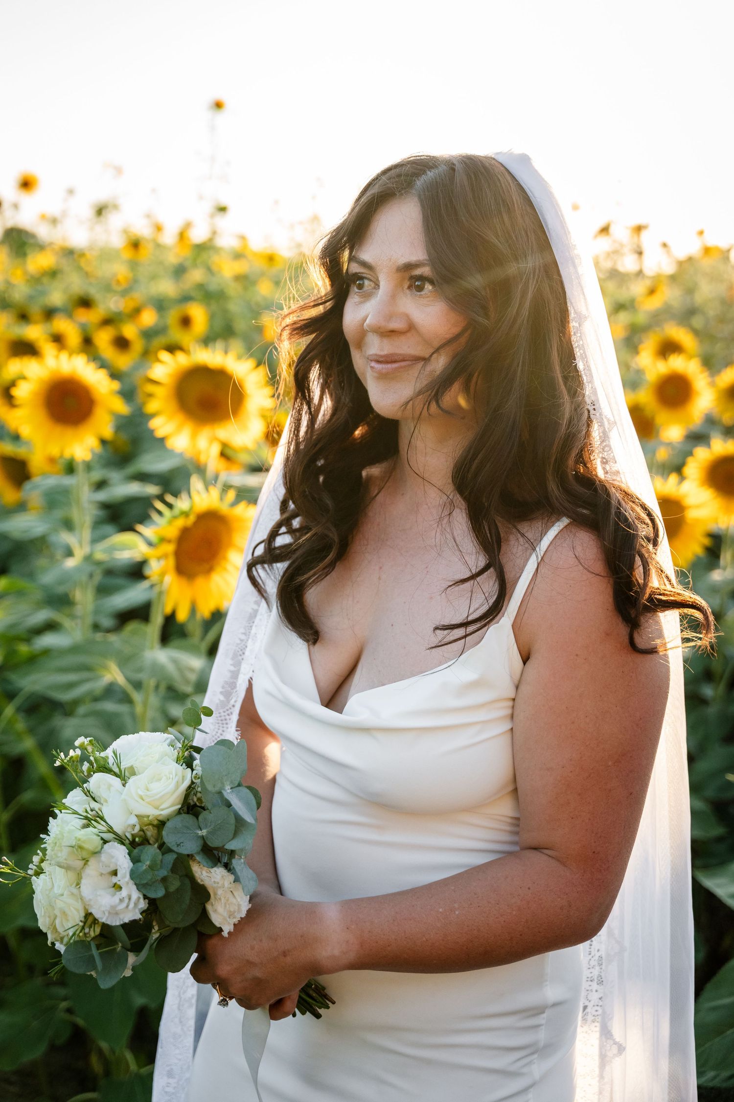 Bride in white gown and veil holds white bouquet in sunny sunflower field.