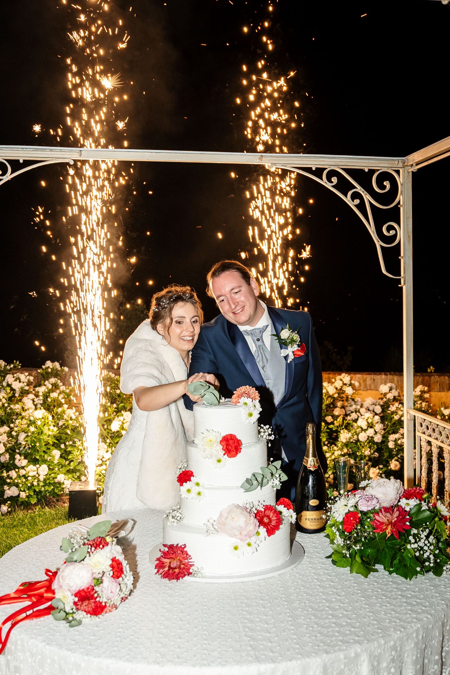 Bride and groom cutting white wedding cake with fireworks display at night outdoor ceremony.