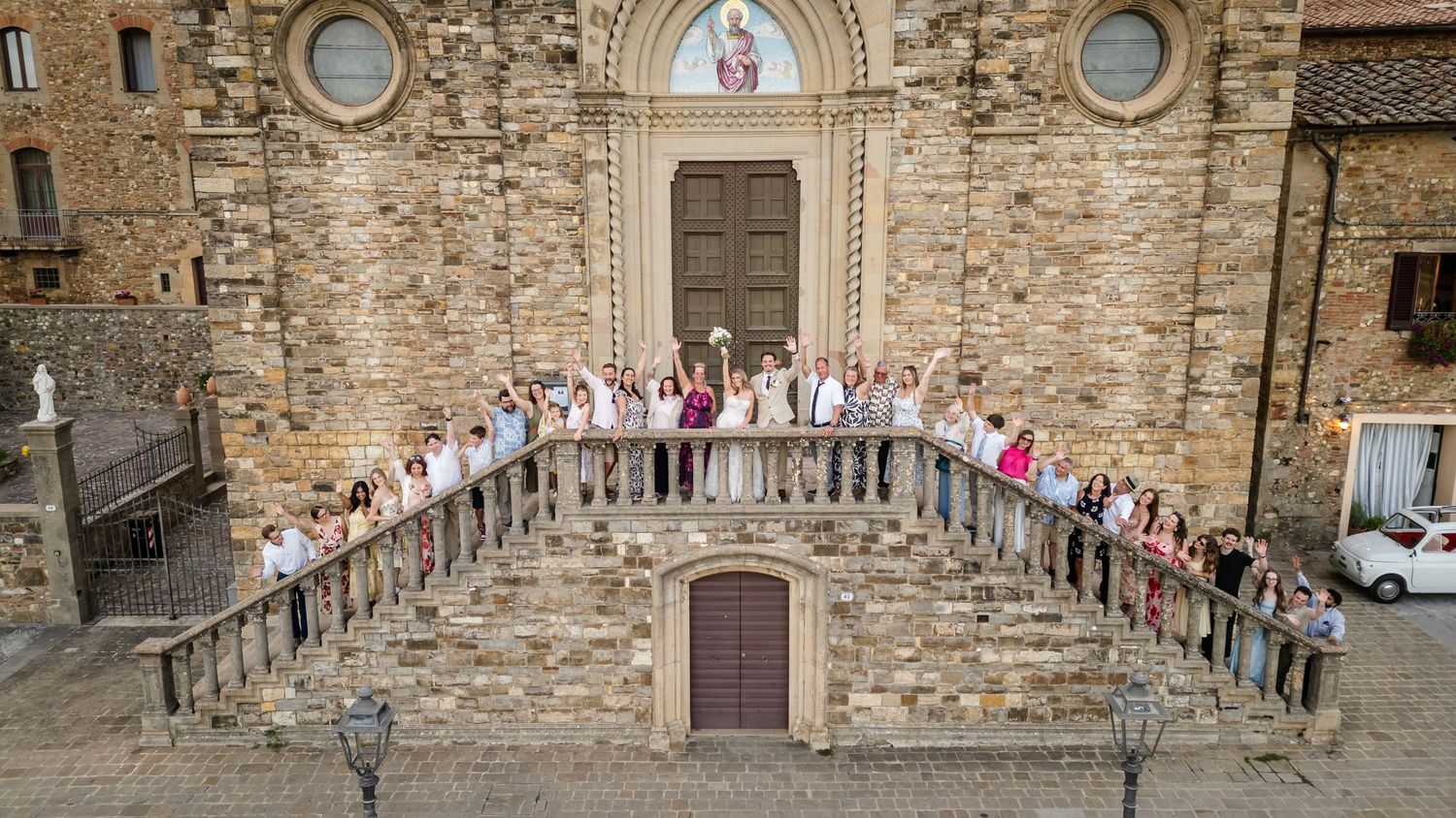 Aerial view of a wedding party posing on stone steps of a historic Italian church in Tuscany.