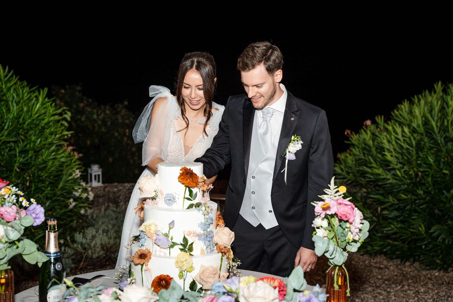 Bride and groom cutting their floral wedding cake outdoors at night, surrounded by colorful blooms.