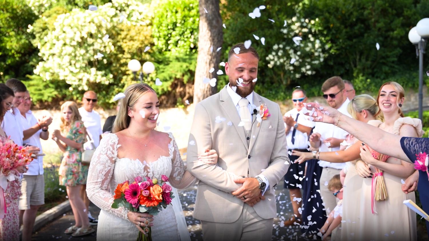 Newlywed couple walks through confetti shower at outdoor wedding ceremony surrounded by cheering guests.