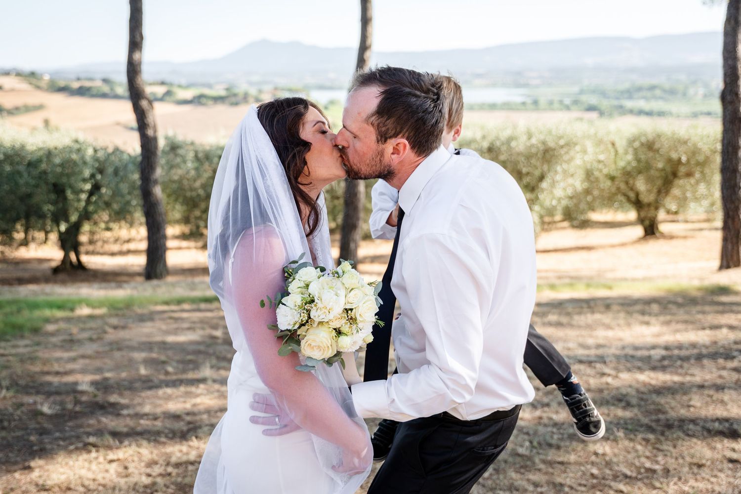 Bride and groom sharing a romantic kiss outdoors, bride holding white bouquet, scenic Tuscan countryside in background.