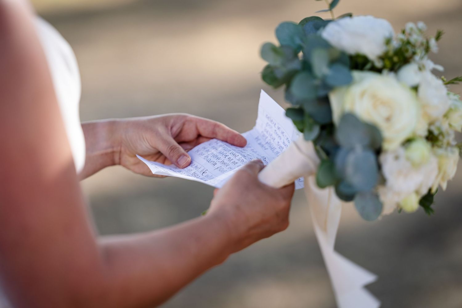 A person's hands hold handwritten wedding vows on white paper with a white floral bouquet in the background.