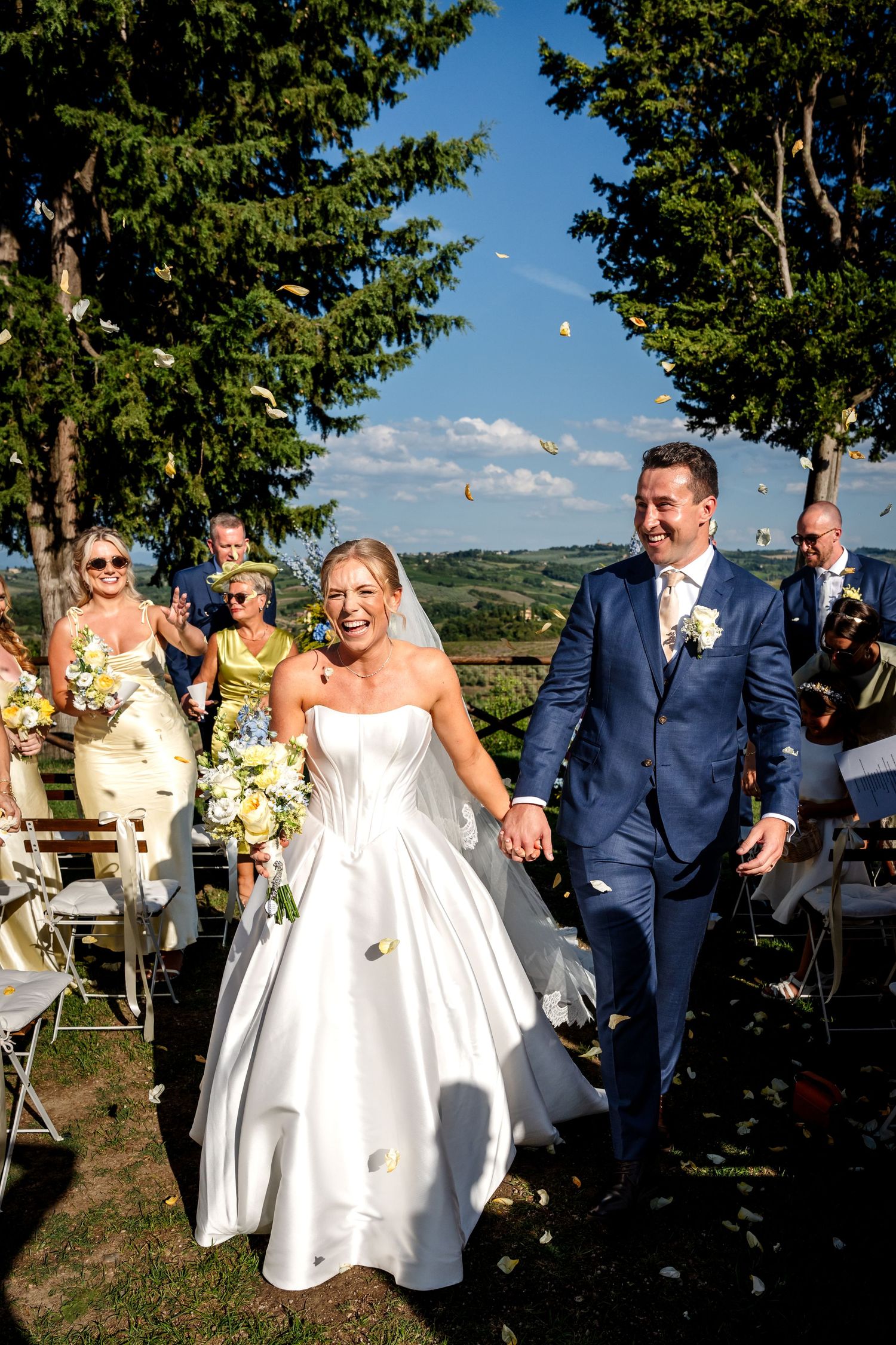 Bride and groom walk down the aisle hand in hand, smiling, with confetti and guests celebrating outdoors.