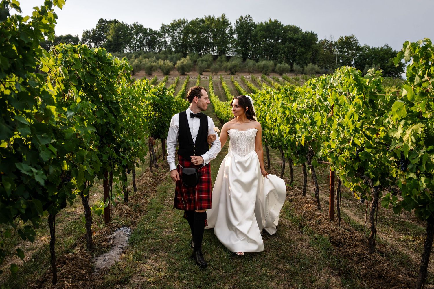 Bride and groom walking through vineyard rows, groom in Scottish kilt, bride in white gown, lush green vines.