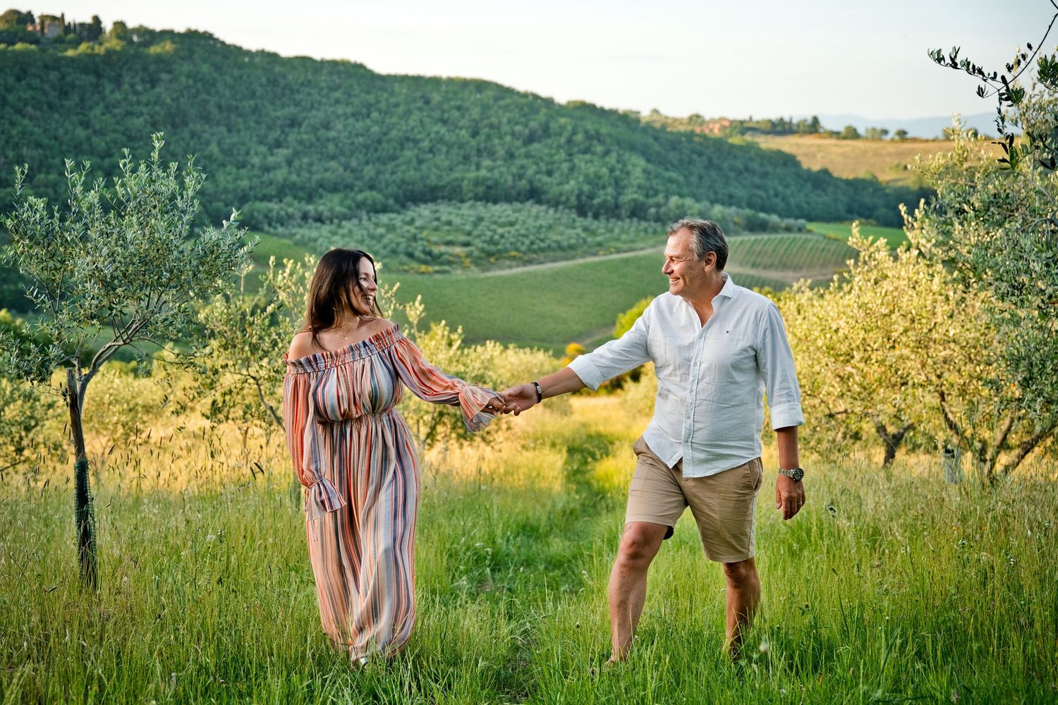 Couple holding hands walking through lush Tuscan countryside with rolling green hills and olive trees.