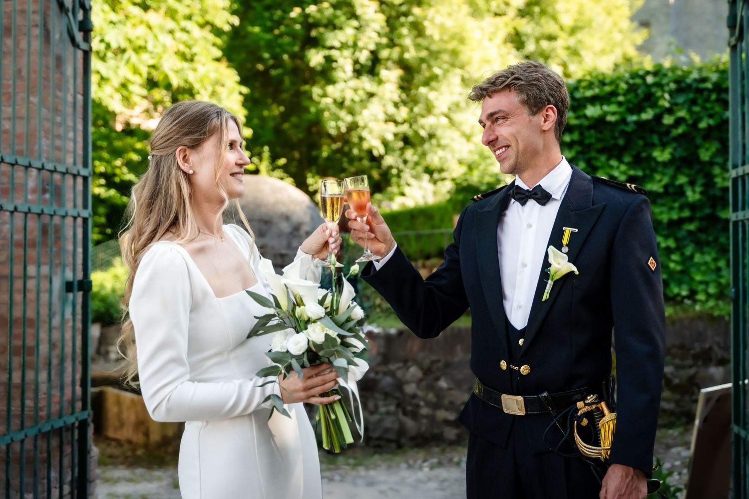 Bride and groom toasting with champagne glasses outdoors, bride holding white bouquet, groom in navy military uniform.