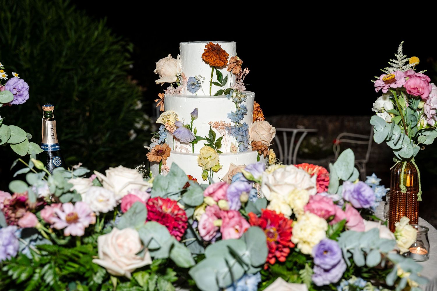Elegant three-tier white wedding cake adorned with colorful floral decorations surrounded by fresh flowers.