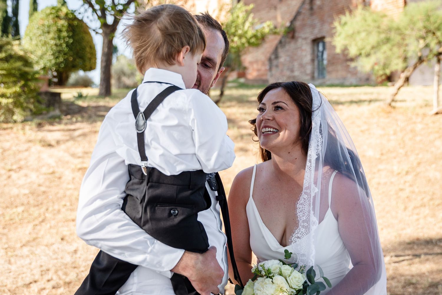 Bride smiles joyfully as groom holds young boy in suspenders at outdoor wedding ceremony.