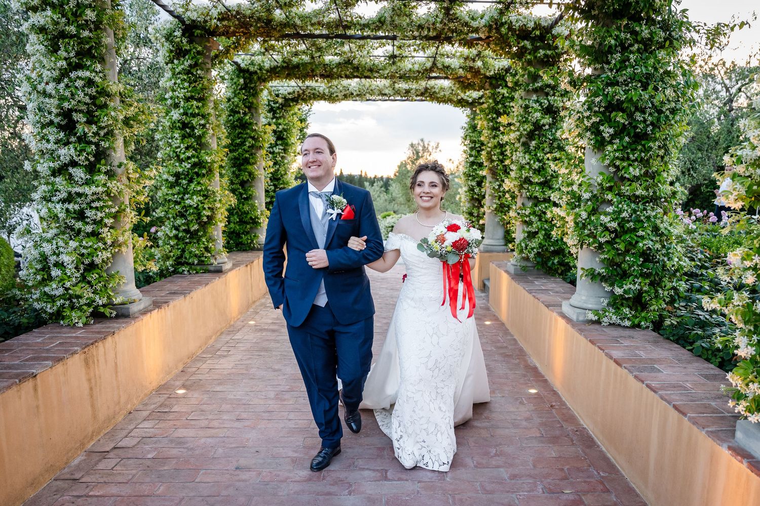 Bride and groom walking under a lush green floral archway tunnel, bride holding red bouquet in elegant white lace gown.