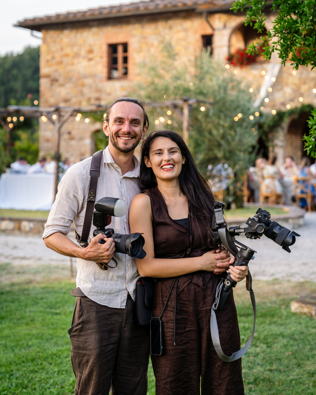 Two photographers smiling and holding cameras at an outdoor wedding venue with string lights and stone building in Tuscany.