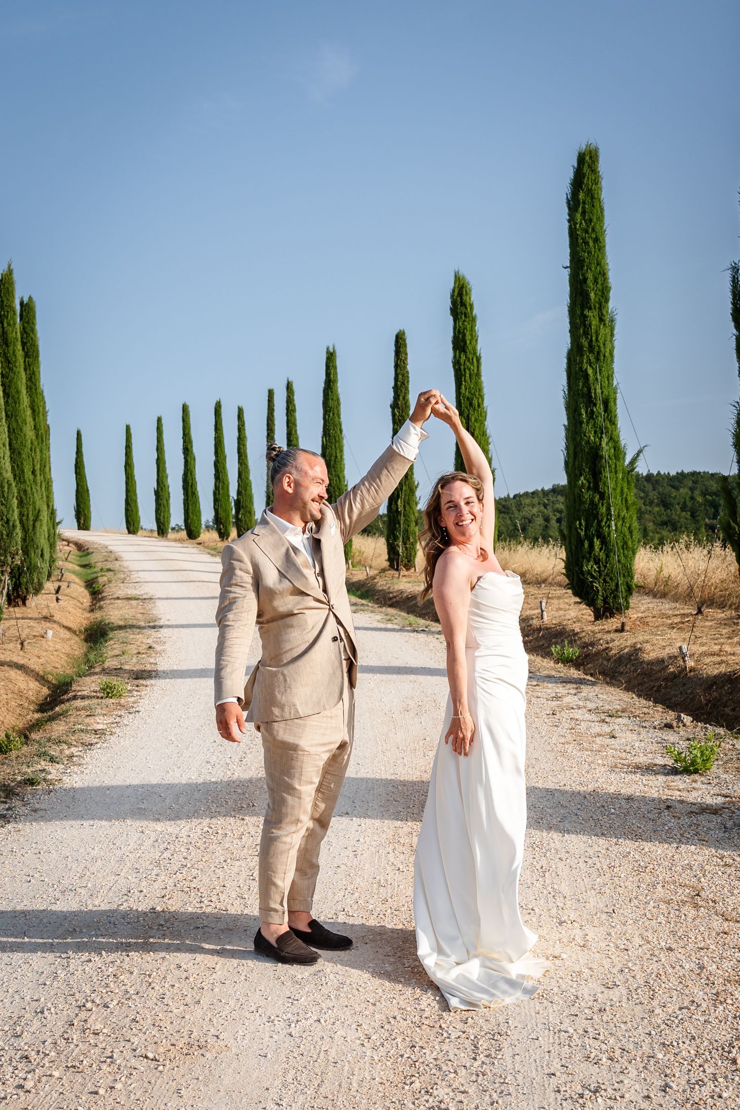 Bride and groom dancing on a scenic Tuscan gravel road lined with tall cypress trees under a clear blue sky.