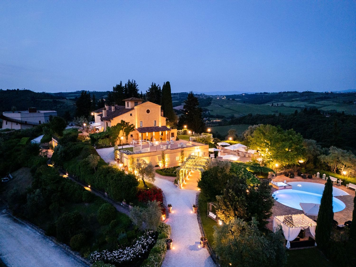 Panoramic twilight view of an illuminated Italian villa resort with swimming pool nestled in rolling Tuscan hills.