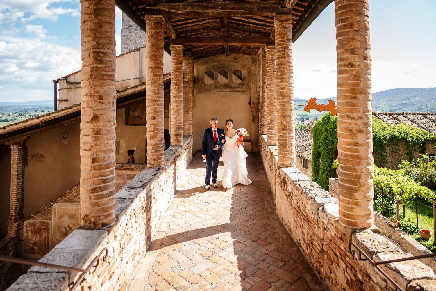 Bride and groom walk along a sunlit stone colonnade terrace with scenic Tuscan countryside views.