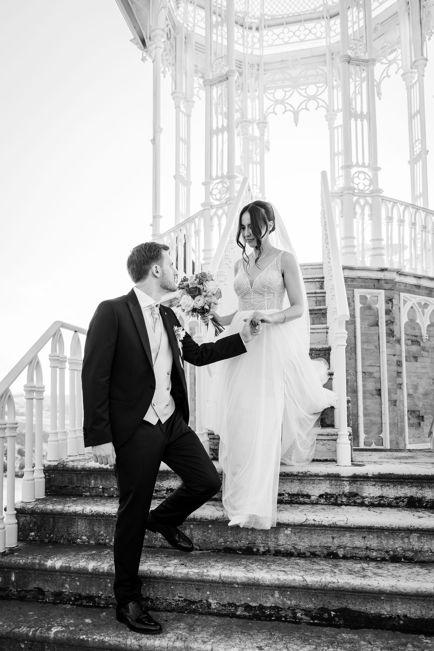 Black and white photo of bride and groom on ornate staircase, groom holding bouquet, elegant wedding portrait.
