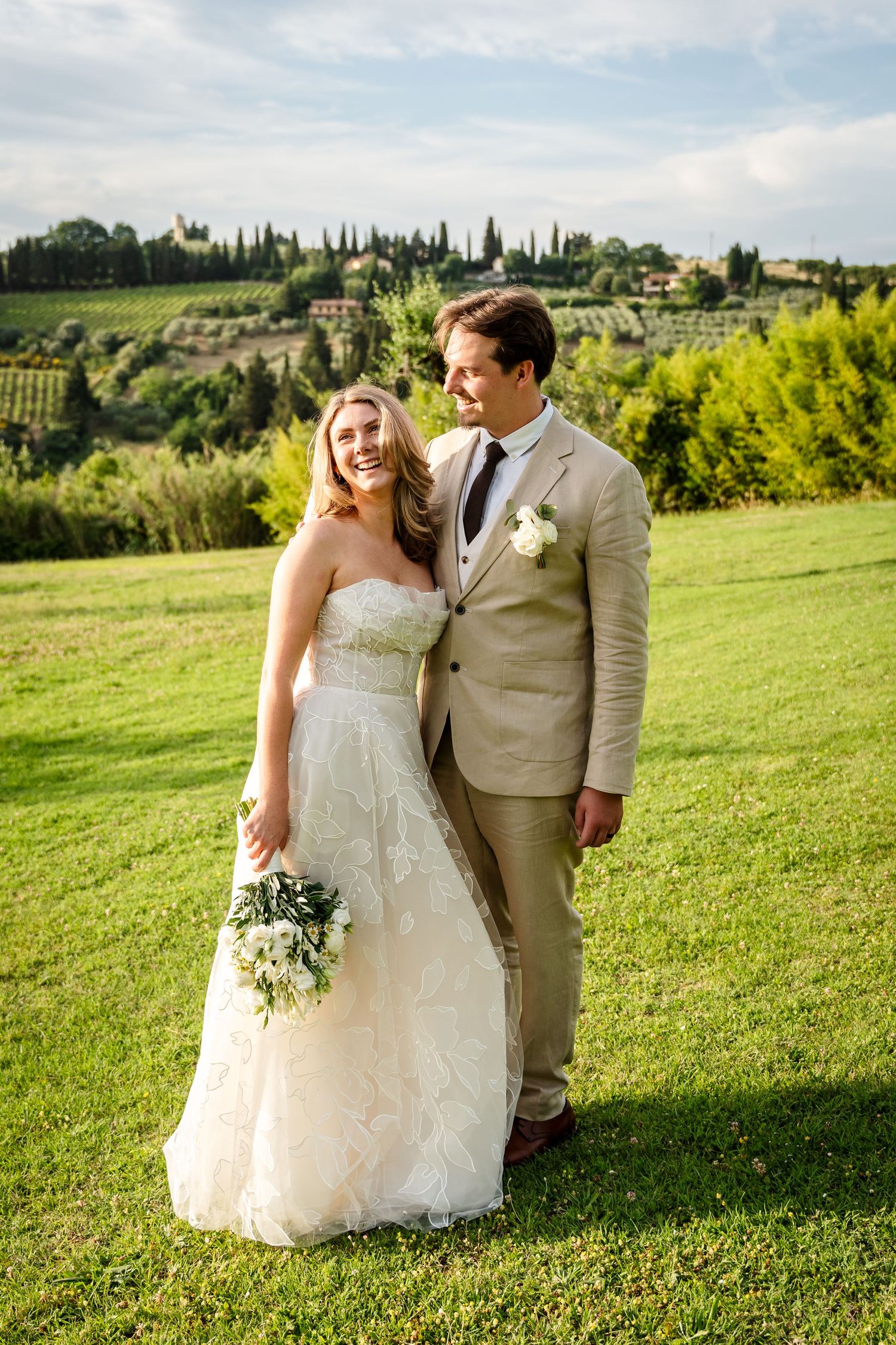 Smiling bride in white gown and groom in tan suit pose on green lawn with Tuscan countryside backdrop.