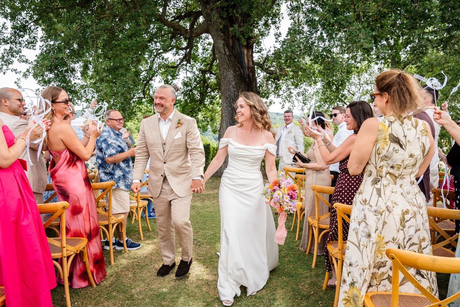 Bride and groom walk down outdoor aisle holding hands while guests celebrate with confetti at sunny garden wedding.