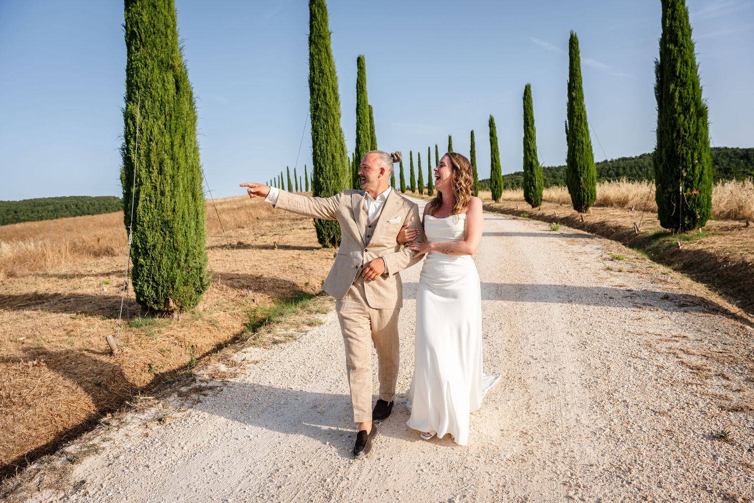 Bride and groom walking on a scenic gravel path lined with cypress trees in Tuscany on their wedding day.