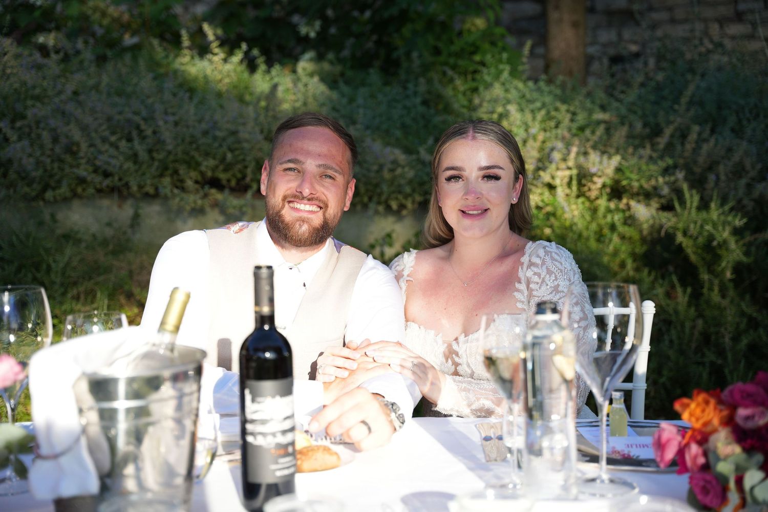 Two people smile at an outdoor dining table with wine glasses and bottles during a sunny afternoon gathering.