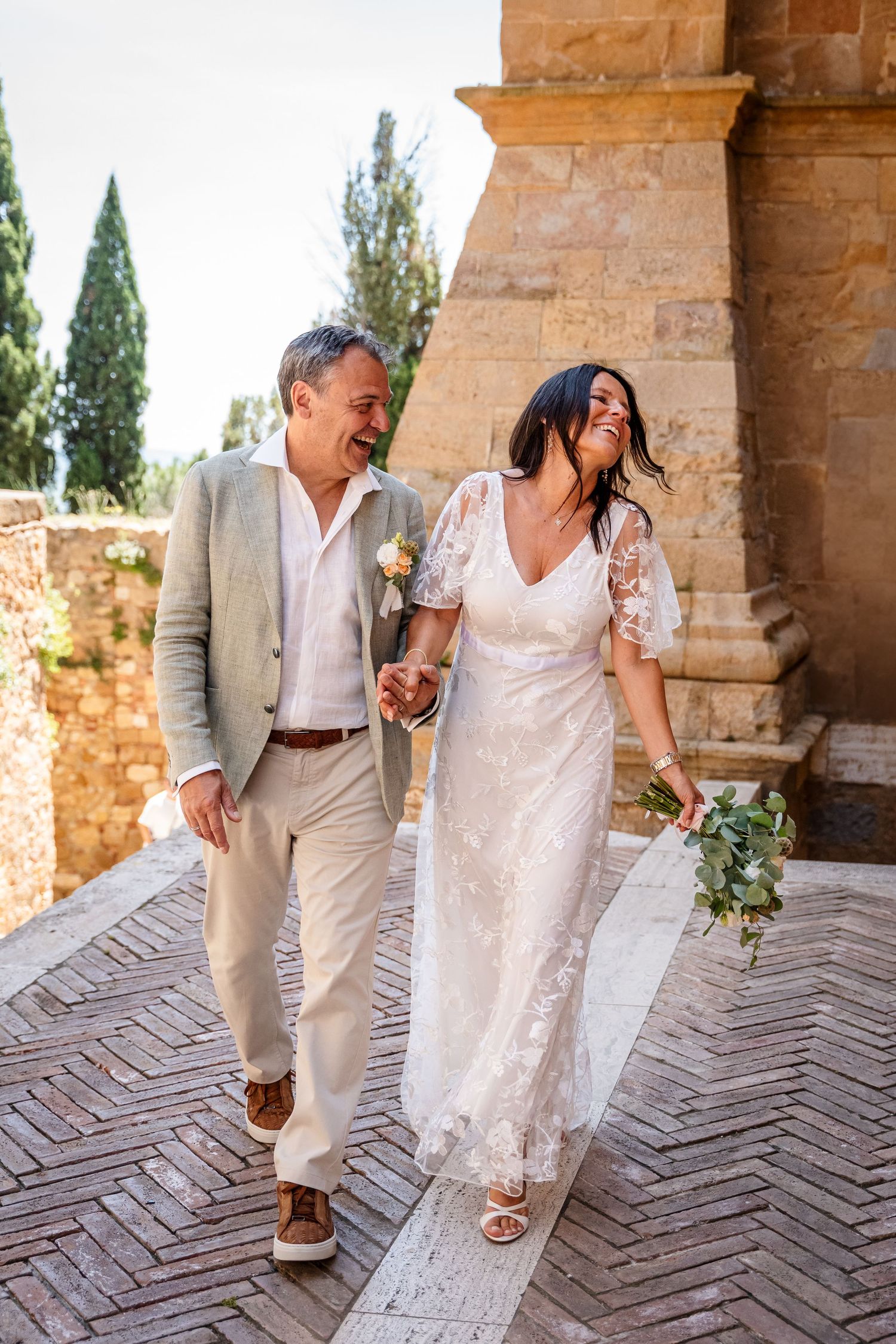 A joyful couple in wedding attire laughs while walking together along a brick pathway at a rustic Italian villa.