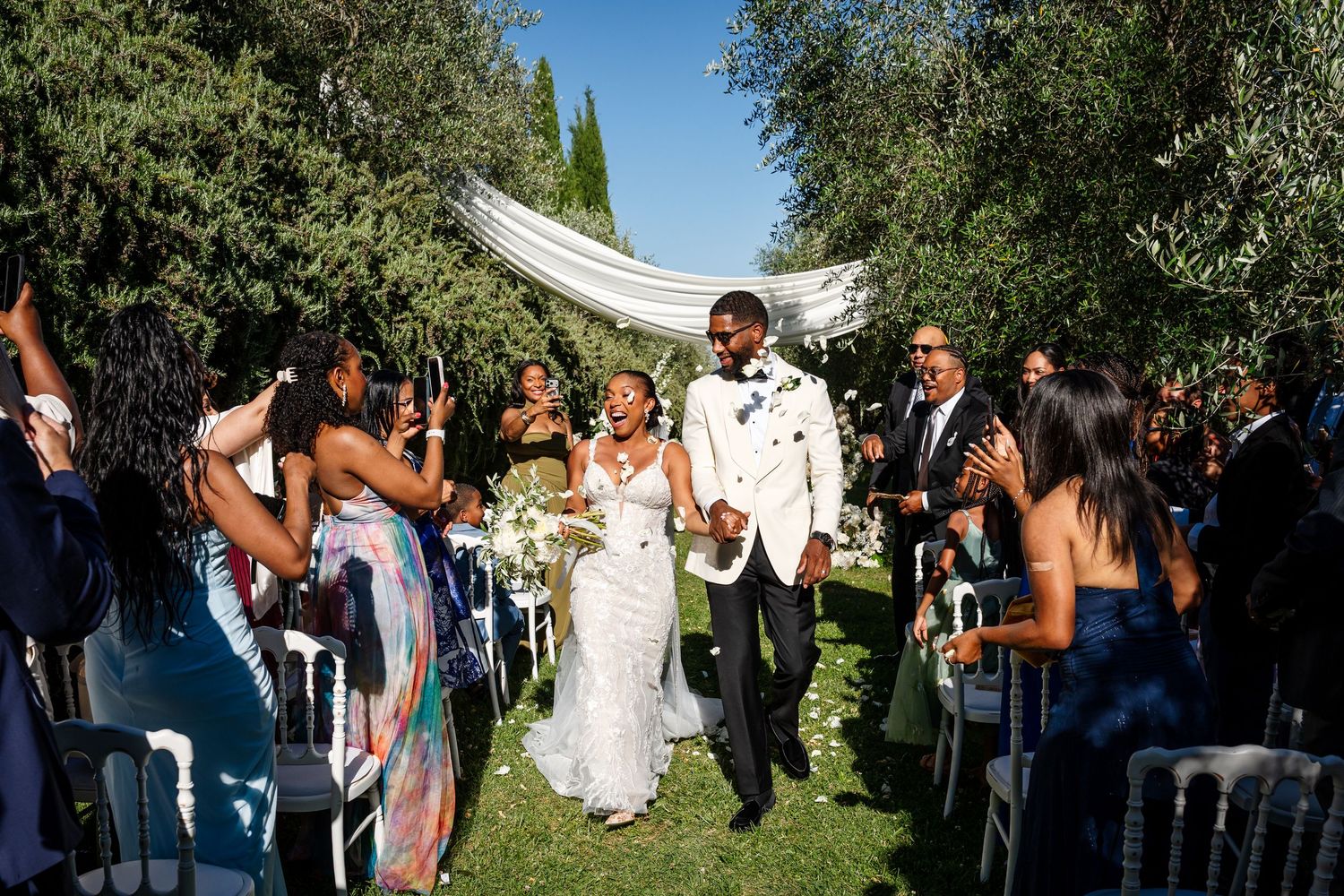 Joyful newlyweds walk down outdoor aisle surrounded by cheering guests at elegant garden wedding ceremony.