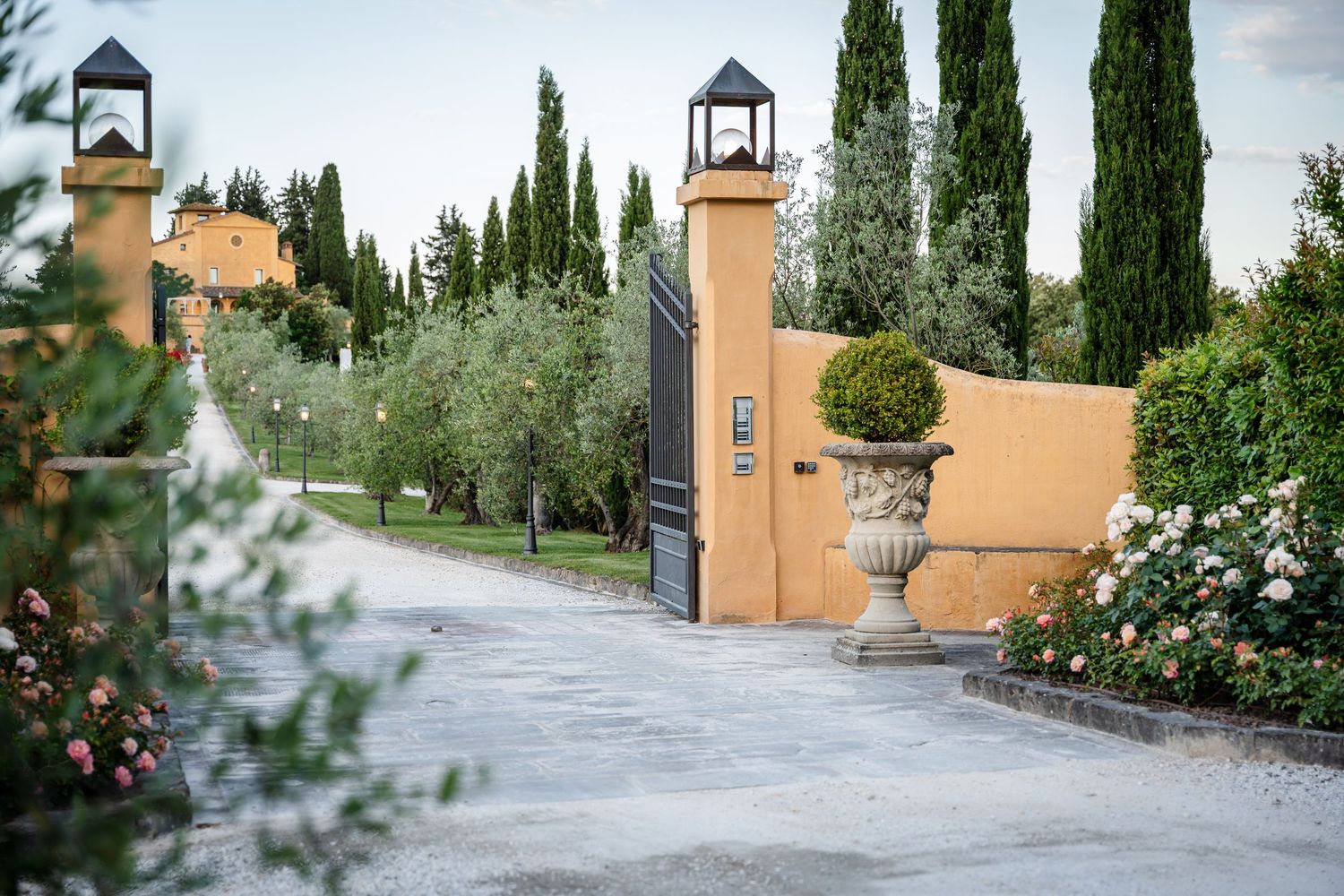 Elegant Tuscan villa entrance with cypress trees, stone urns, and ornate iron gates at dusk.