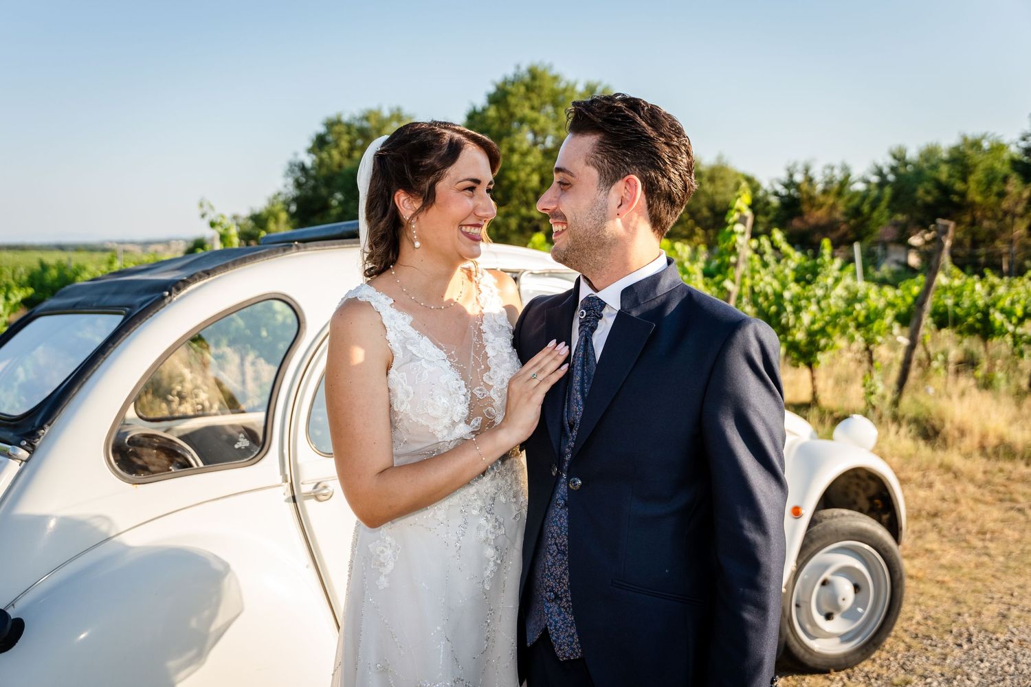 A bride and groom smile at each other beside a vintage white car surrounded by lush green vineyards on a sunny day.