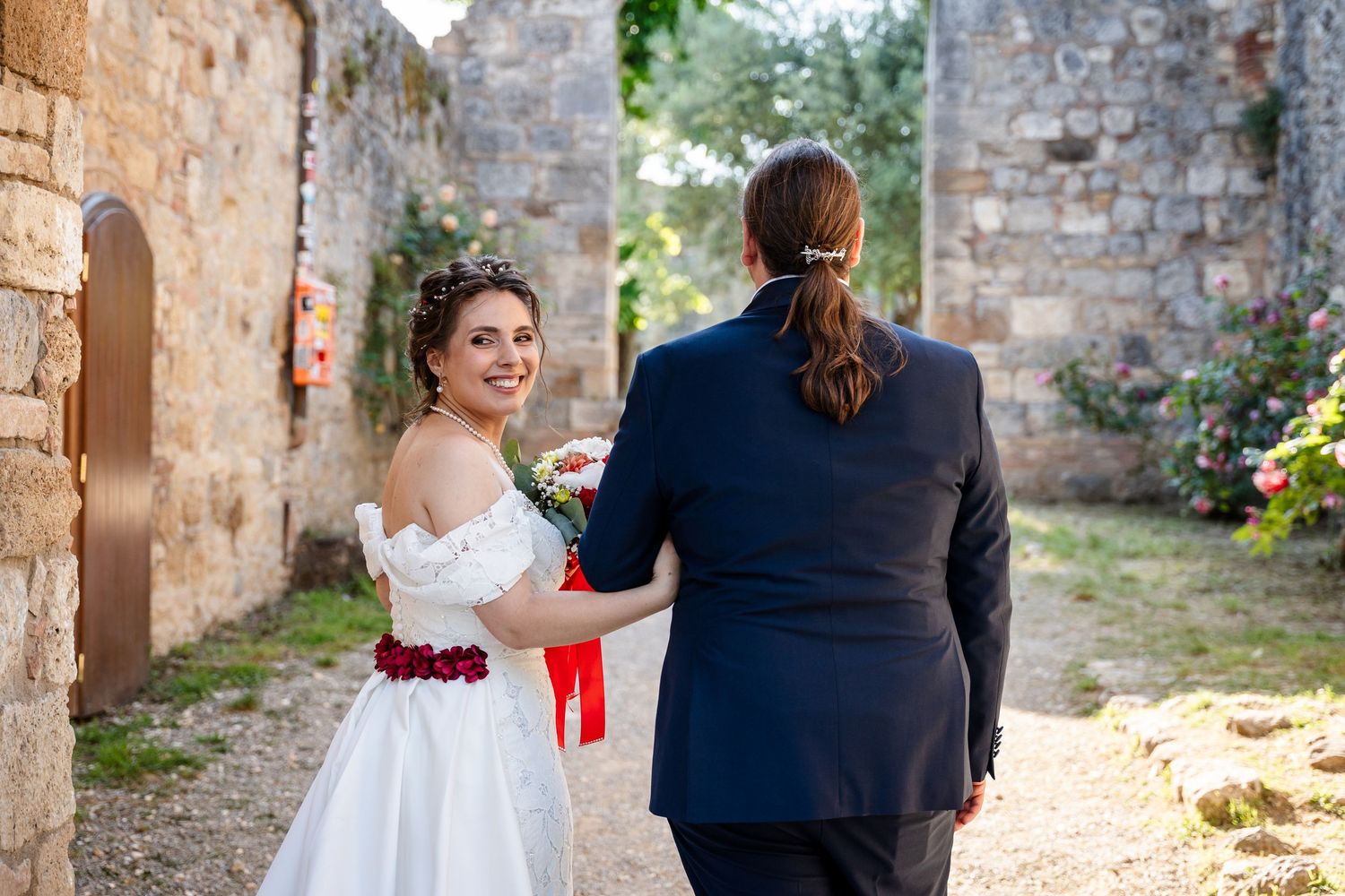 Bride in white off-shoulder gown holds groom's hand walking through stone ruins on wedding day.