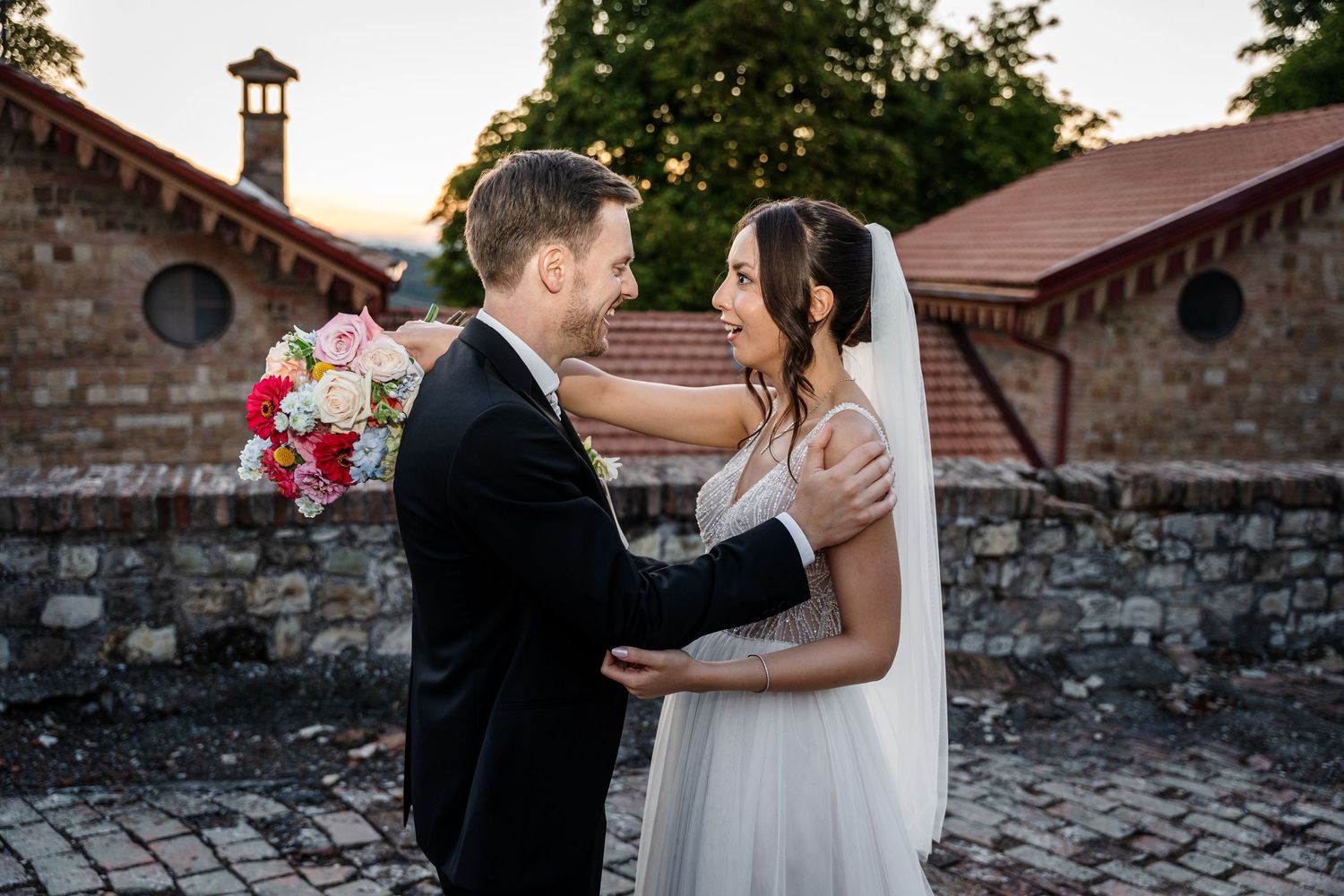 Bride and groom share a romantic moment outdoors at sunset, with bride holding colorful bouquet near stone building.