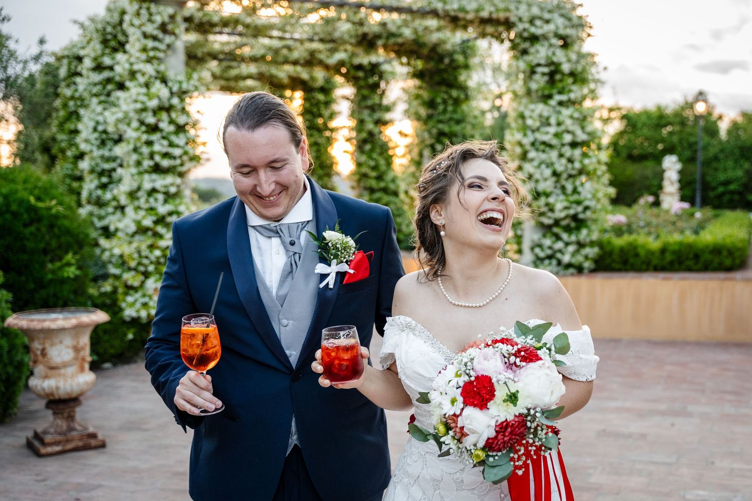A joyful couple shares a laugh while holding cocktails at their outdoor garden wedding celebration.