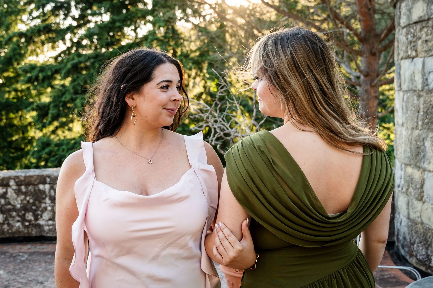 Two women in elegant dresses, pink and olive green, share a moment outdoors near stone wall and lush trees.