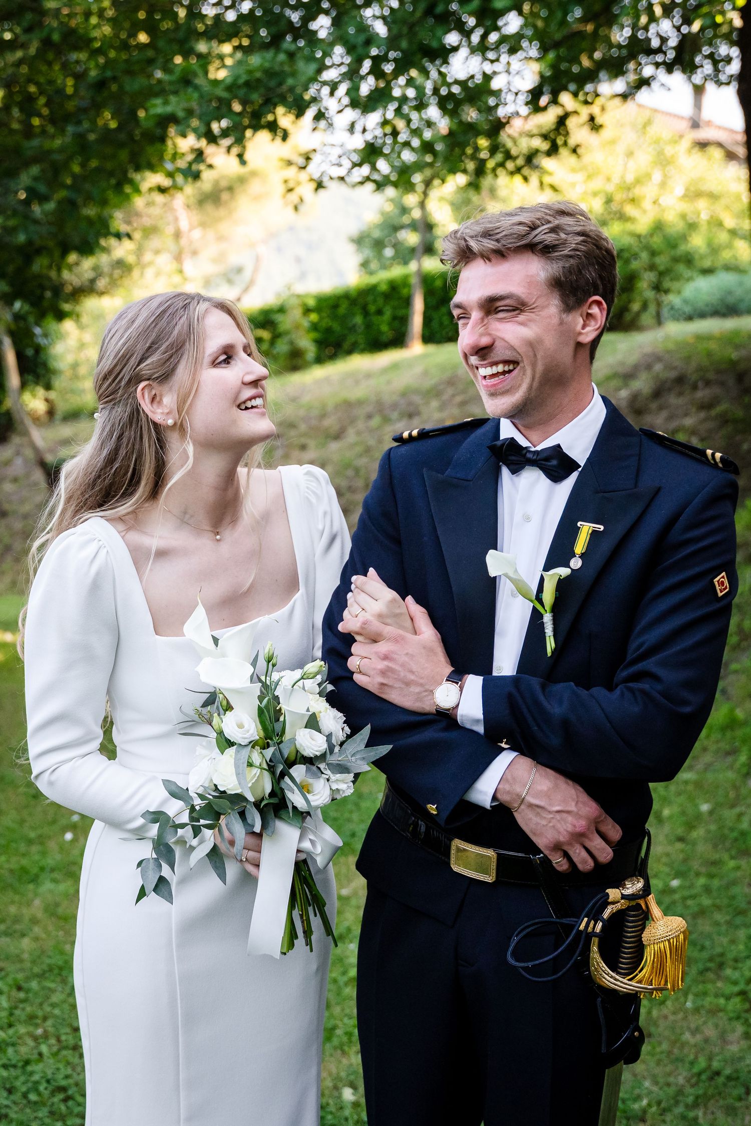 Happy newlyweds smile outdoors, bride in white gown holding bouquet, groom in navy military suit with bow tie.
