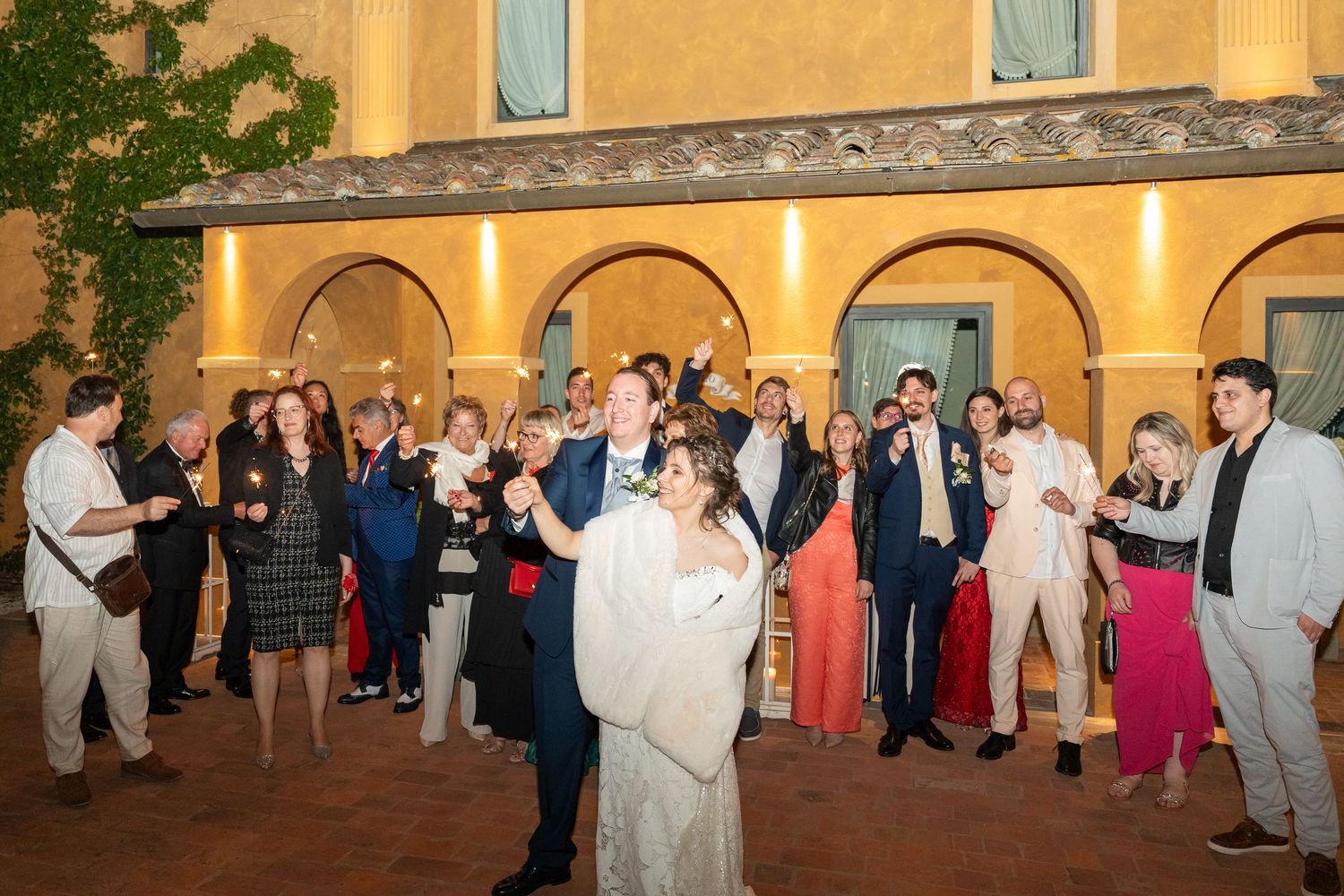 Newlyweds dance as wedding guests celebrate with sparklers in a romantic Italian courtyard at night.