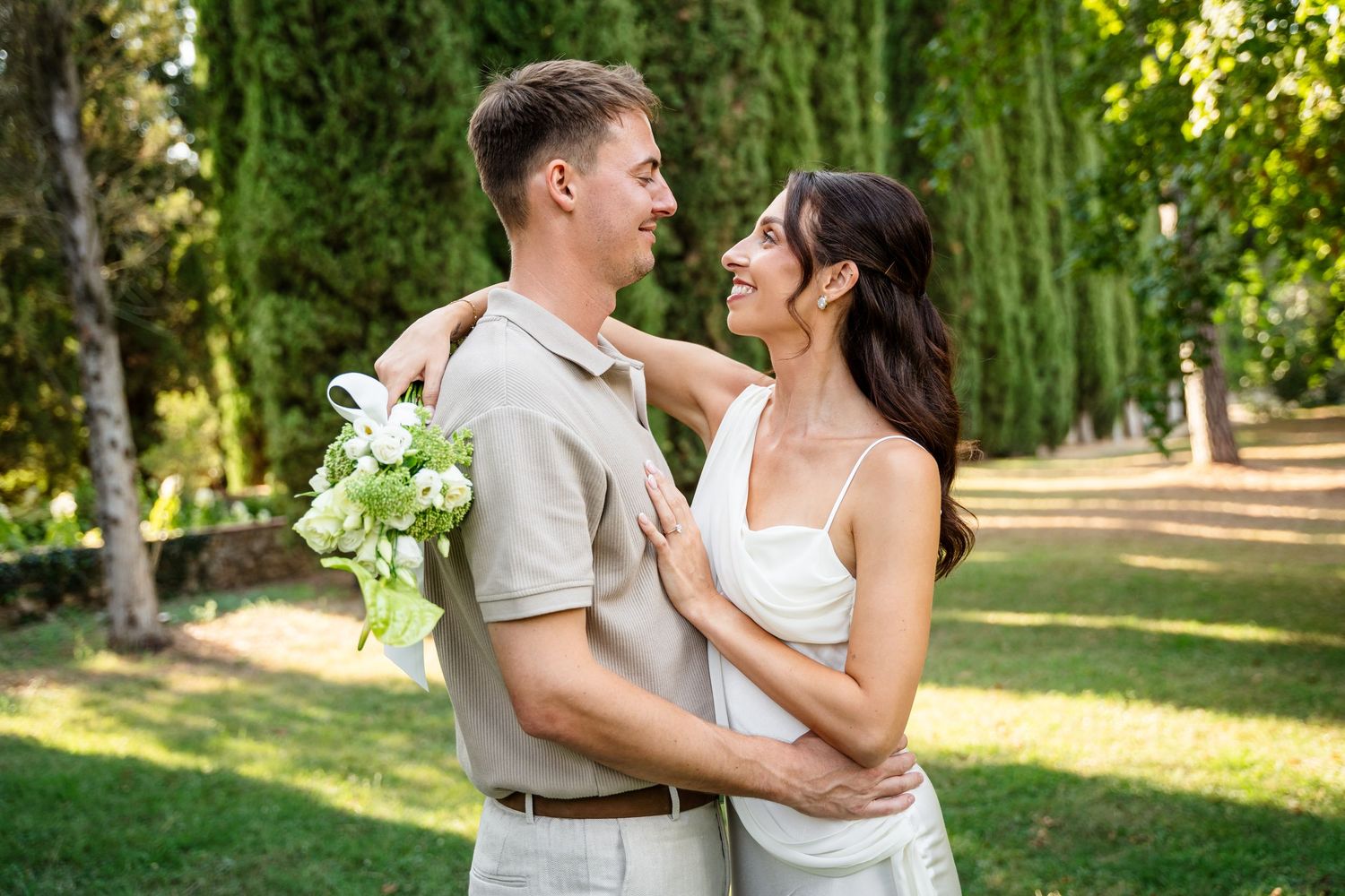 Happy couple embracing in a green park, man holding white flowers, woman in white dress smiling.