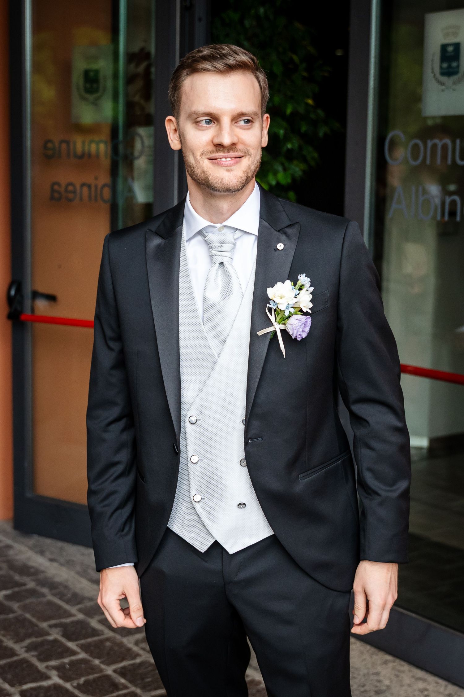 Smiling groom in black tuxedo with white vest and floral boutonniere standing outside a building.