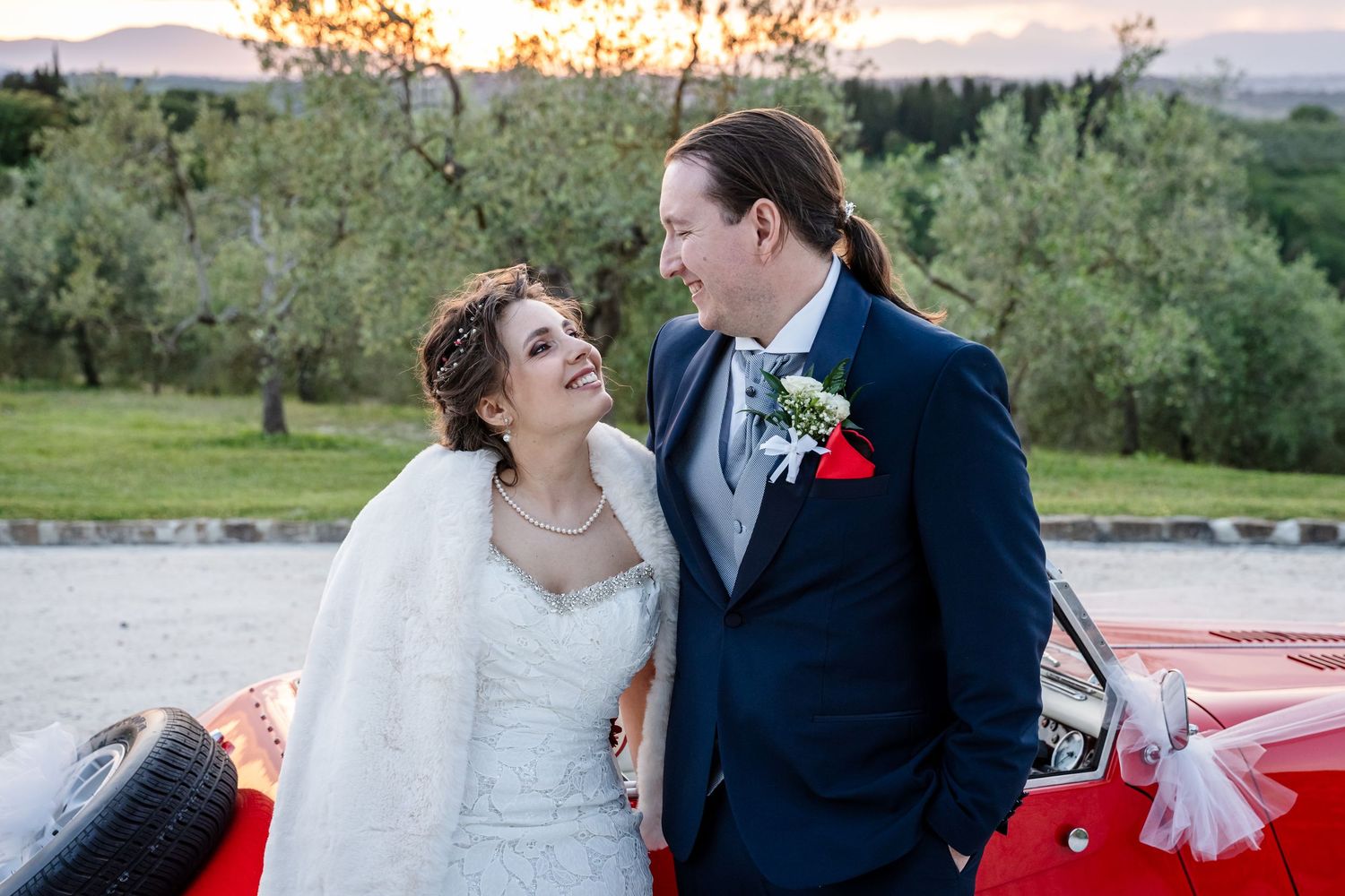 Newlywed couple sharing romantic moment beside red vintage car at sunset in Tuscan countryside.